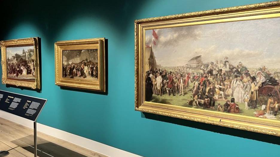 Three paintings in gold frames from the Victorian era feature crowds of people with children sitting on the grass. The middle painting appears to be in a railway station with a high roof.