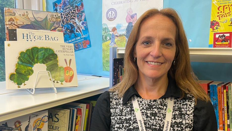 A teacher stands by books in the special school's library. She has brown hair and a black and white top on. 