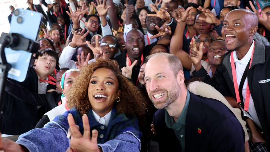 Prince William and Nomzamo Mbatha pose for a selfie with young people during the Earthshot Prize Climate Leaders Youth Programme in Cape Town, South Africa. Both are smiling and are surrounded by dozens of smiling young people.