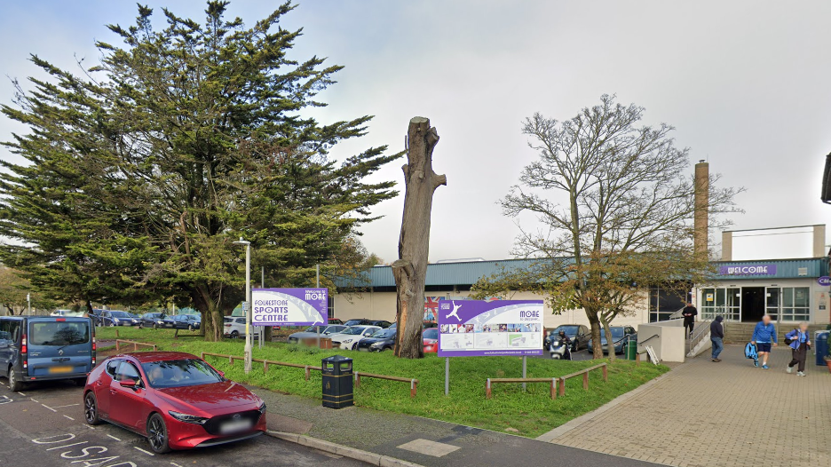 The front of Folkestone Leisure Centre, a grey building with purple signage. The leisure centre has a grassy verge in front of it as well as a large tree and cars parked in front of it.