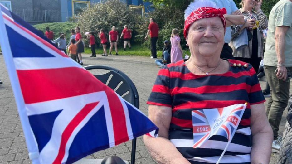 Donna Thompson is in a striped red and black top and wearing a red hairband.
She is standing beside a large Union flag and has another in her hand.
A number of children are playing in the background