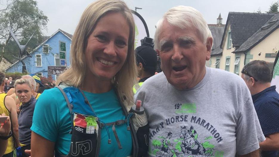 BBC news prestner Sophie Raworth with Mr Green at one of the races, she is wearing a blue top with medium length blonde hair. Mr Green is in a grey tshirt with short white hair.