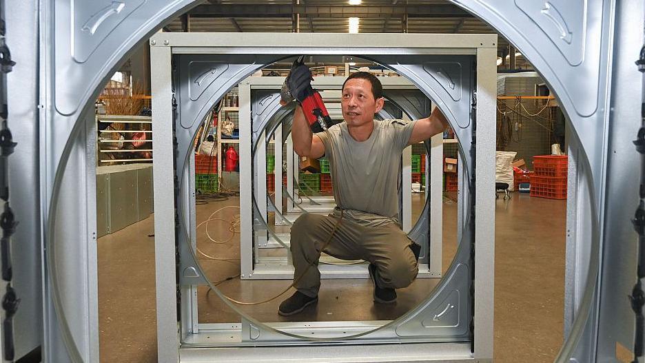 An employee at a wind turbiner manufacturing company uses work tools to put together some steel parts. In the image, he is pictured squatting at the centre of one of the steel frames. 