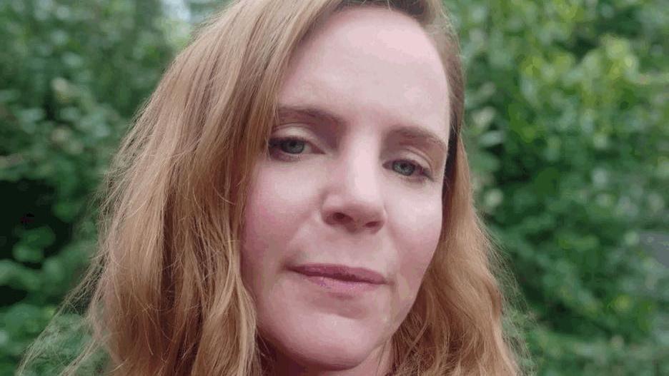 Holly Sandiford has long auburn hair, and is looking directly at the camera. She is standing near some trees, which are in soft focus.