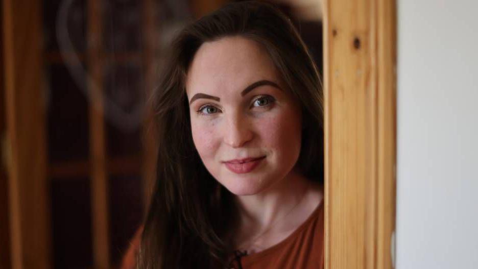 Sara Spencer standing next to a door frame. She has long brown hair and is slightly smiling at the camera.