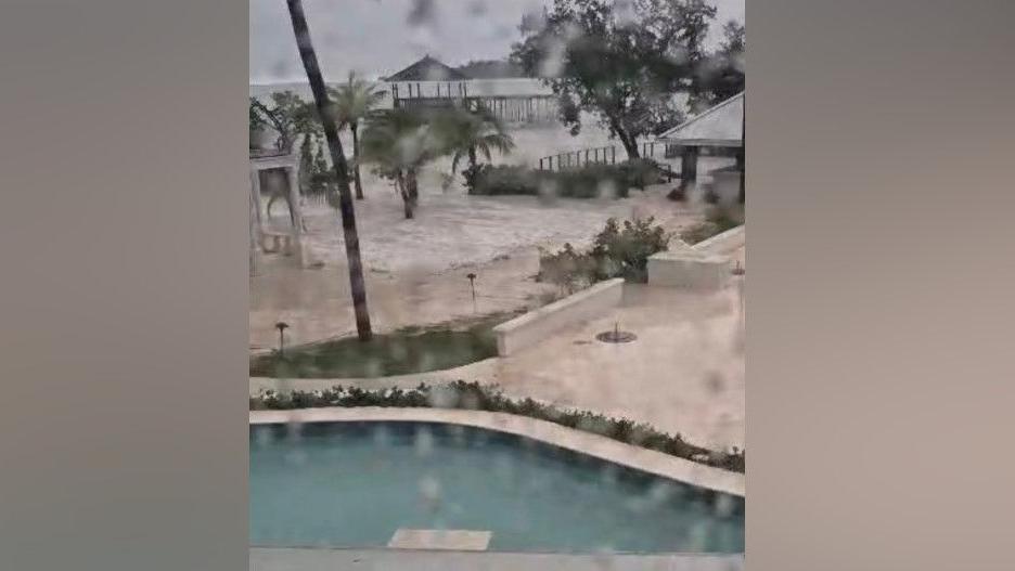 View out of a rain-spattered window at the grounds of a hotel. Part of a swimming pool can be seen in the foreground, with palm trees, shelters, and the sea behind it.