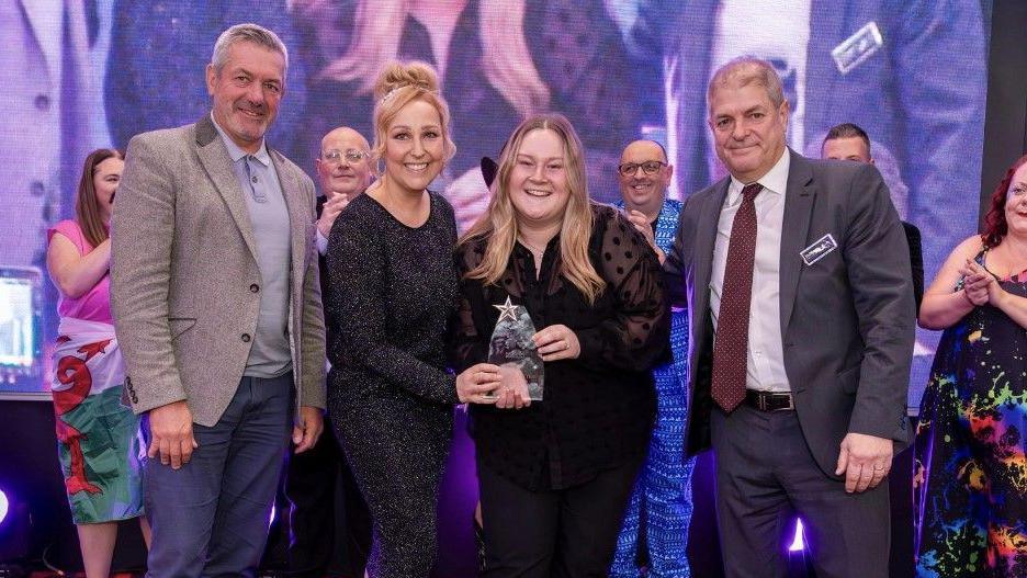 A group of people stand in a line presenting Ms Hunt with her award. They are smiling at the camera while Ms Hunt is handed her trophy. Other people can be seen behind them smiling and clapping. 