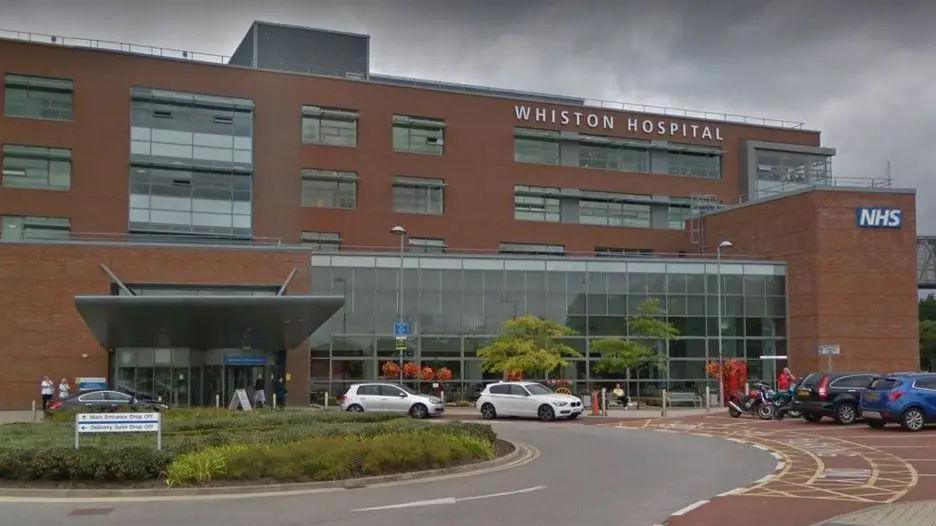 Exterior view of Whiston Hospital showing main entrance with a roundabout in front and a number of cars parked