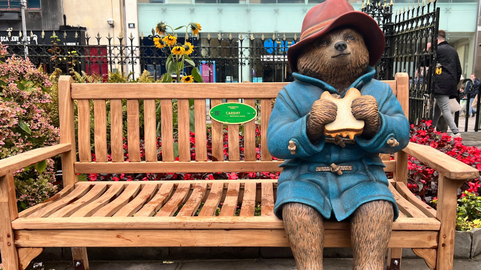 Paddington Bear statue in a blue duffle coat and red hat, eating a jam sandwich on a bench in St John's Gardens, Cardiff. 