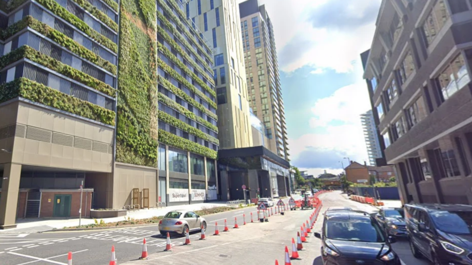  A Google Maps image of Victoria Way in Woking. There is a large hotel development building to the left , white and orange cones in the centre and cars on the right. 