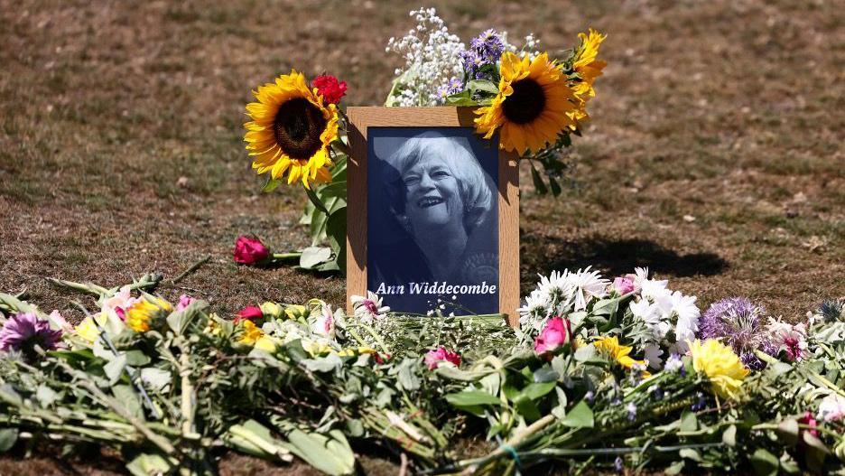 A framed black and white picture of Ann Widdecombe smiling is placed upon a grassy area, surrounded by flowers including sunflowers