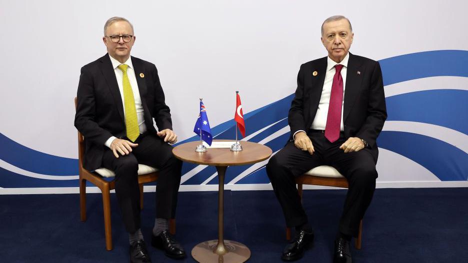 Two men in suits sit in chairs. Between them is a round table with two small flags: one Australian and the other Turkish