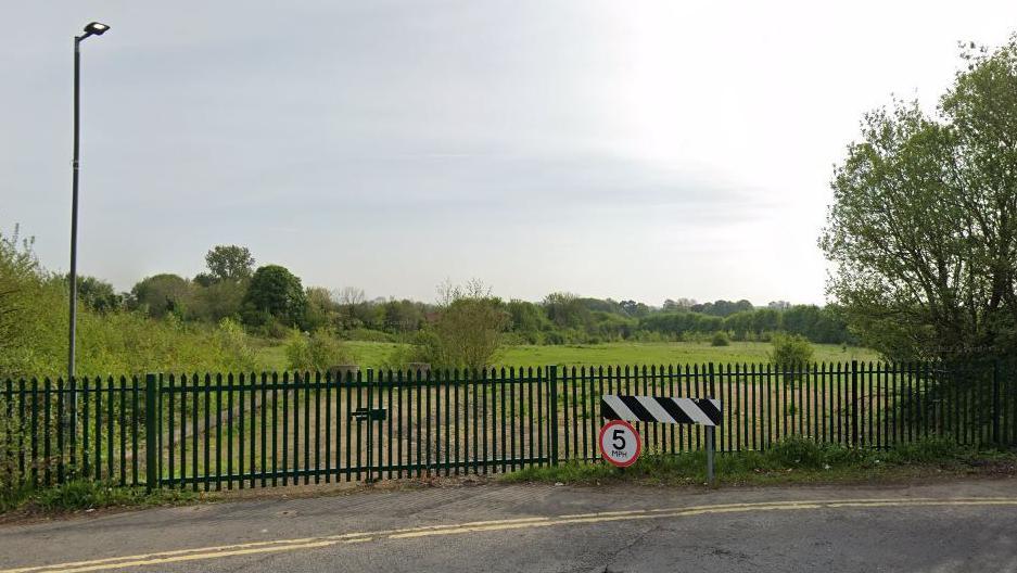 A metal gated fence next to a 5mp sign stands in front of a grass field with trees in the background.