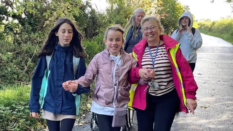 Three girls and two women walking down a country lane, with green trees and bushes to either side. In the front row, a girl has long brown hair, a blue jacket and black leggings. She is holding hands with a second girl who has blonde hair, a pink jacket and black leggings. Her other hand is held by a woman who has blonde hair, glasses, pink jacket, striped top, lanyard and dark trousers. In the back row a woman with long blonde hair and a dark top walks with a girl who has long blonde hair, a grey hoody and dark trousers.