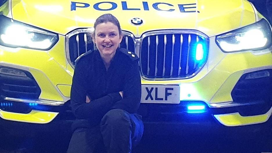 A woman in a police uniform crouches with her arms crossed and smiling in front of a police car. 