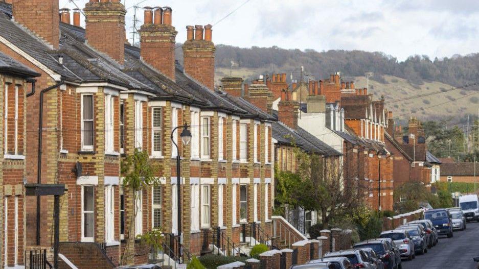 A row of redbrick terraced housing. There is countryside in the background and cars are parked along the street.