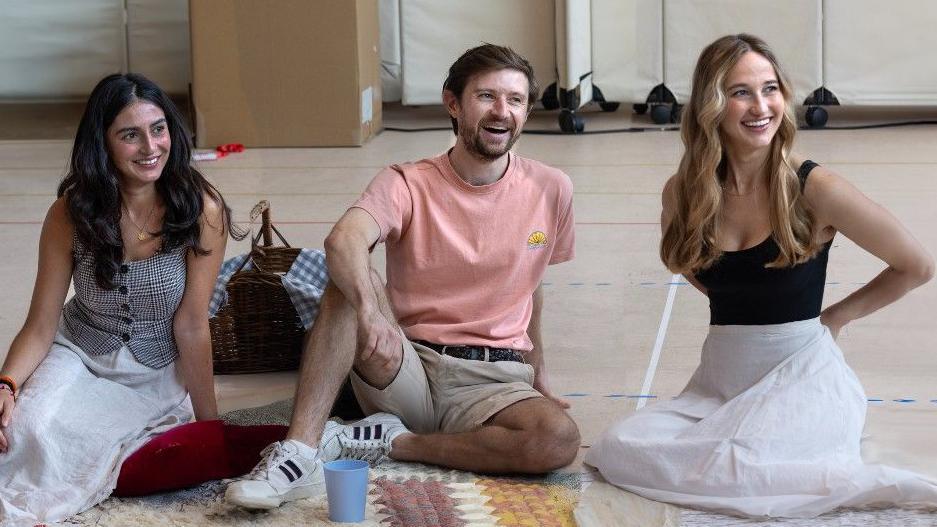 Three actors are sitting on the floor. Maiya Louise Thapar has long dark hair and is smiling. Peter Losasso is a bearded man wearing a T-shirt and shorts. India Shaw-Smith has long tousled blonde hair and wears a black vest and long white skirt. They are all laughing and looking slightly off camera.