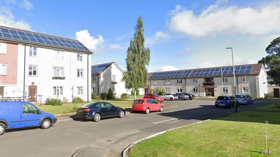 A photos of the Barlow Road housing estate showing a mowed lawn, several cars parked on the road and blocks of flats. The sky is blue with some clouds.