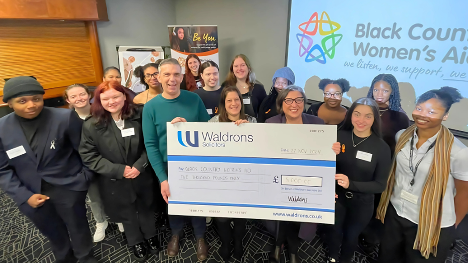 A group of people all smiling and standing around a large blue and white cheque that says "Waldrons solicitors" in the top left corner. The group include several women and a couple of men. They are indoors and stand in front of a screen showing the Black Country Women's Aid logo and text of their title.