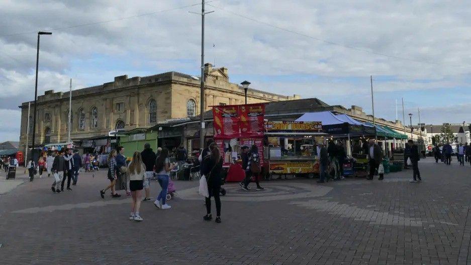 Shoppers mill about around a town centre market. Some food stalls, cast in shadow, are dotted about. A large civic building looms overhead.