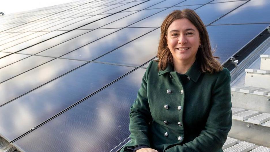 Alex Mayer is sitting on white steps next to a roof or platform covered in solar panels. She is smiling a the camera and is wearing a dark green coat with double breasted silver buttons. She has medium length brown hair.