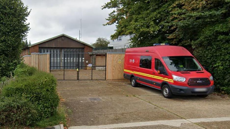 Google Street view of the old fire station. The site is sealed off with wooden panels and padlocked metal gates. On the driveway in front of the gates is a red fire service crew van.