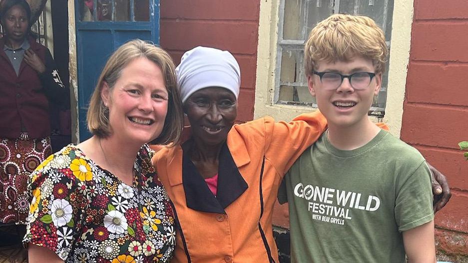 Gill, who has short brown hair, and her son Sam, who has thick fair hair and glasses, pose for a picture with a Kenyan lady whose hair is covered with a lilac shawl.