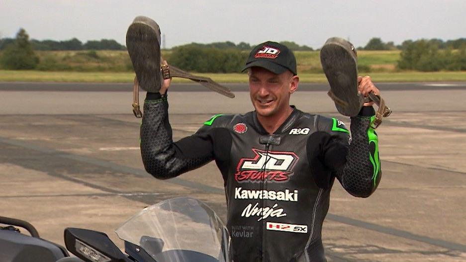 Jonny Davies in black motorcycle gear, holding up his stunt shoes on the airfield at Elvington