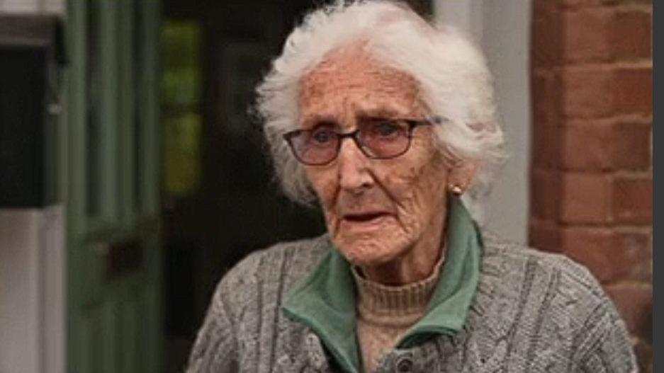 Elizabeth Stone with medium-length white hair and brown-framed glasses, wearing a grey sweater over a green fleece. There is a house door and brick wall behind her.
