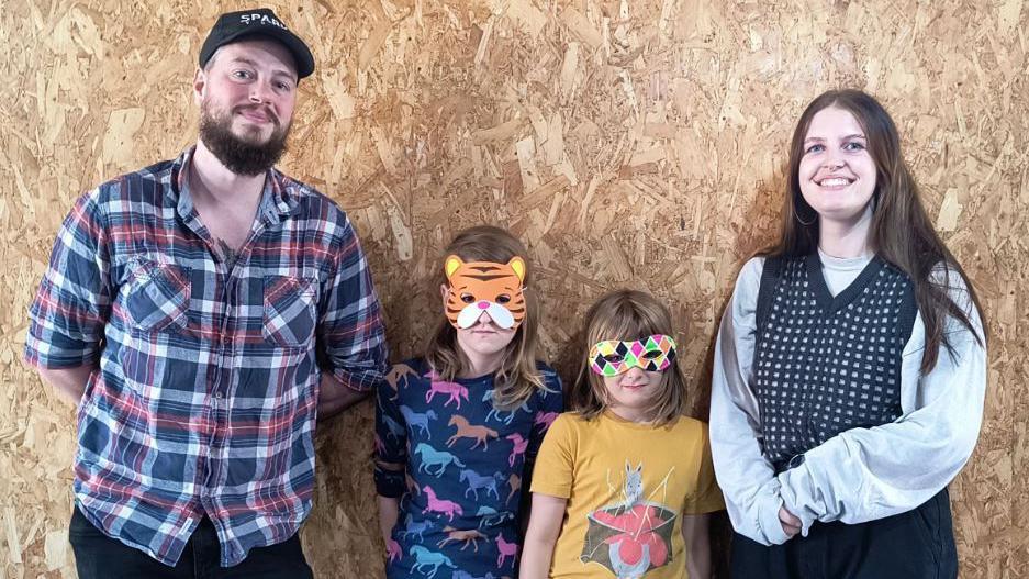 Two Spark staff - Gemma Spivey and Matthew Kirk - meet Ada and Mabel from YorKAOS - all are pictured standing by a chipboard wall, the children wearing bright coloured masks.