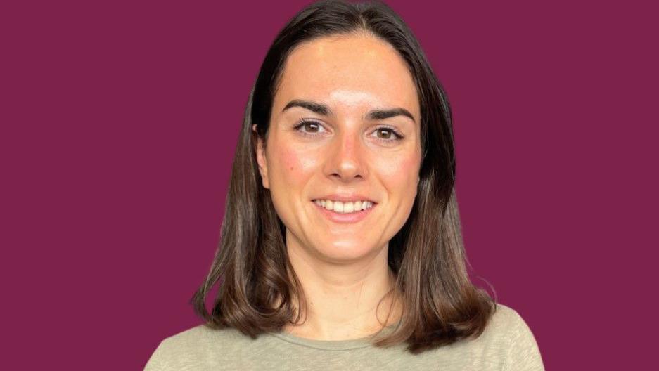 Abigail, who has shoulder length brown hair, smiles at the camera. She is wearing a beige top and standing in front of a burgundy background. 
