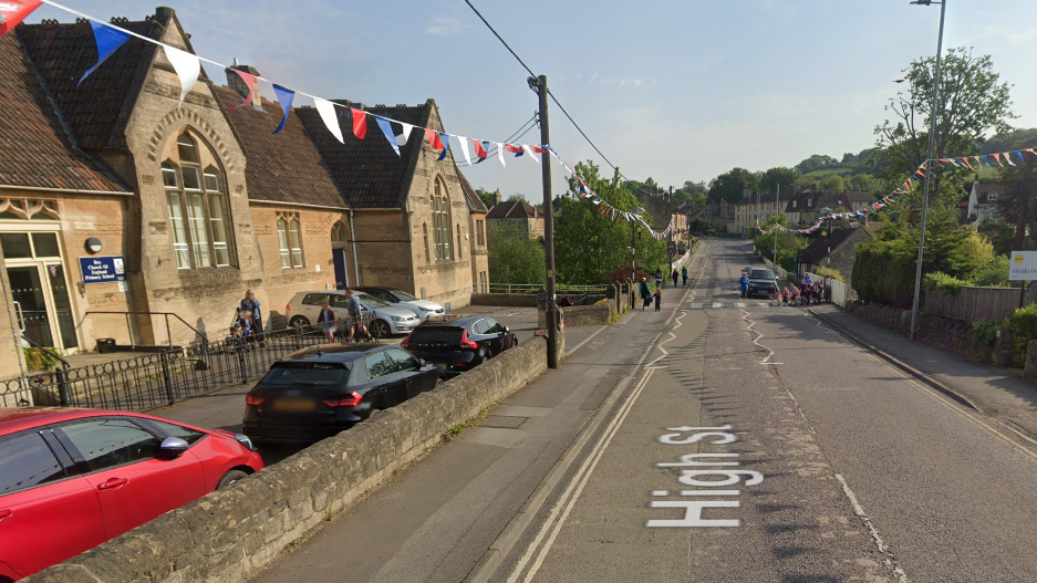 Box Primary School building to the left with a road on the right and zebra crossing in the distance with a group of people about to cross. There is bunting hanging down the road.