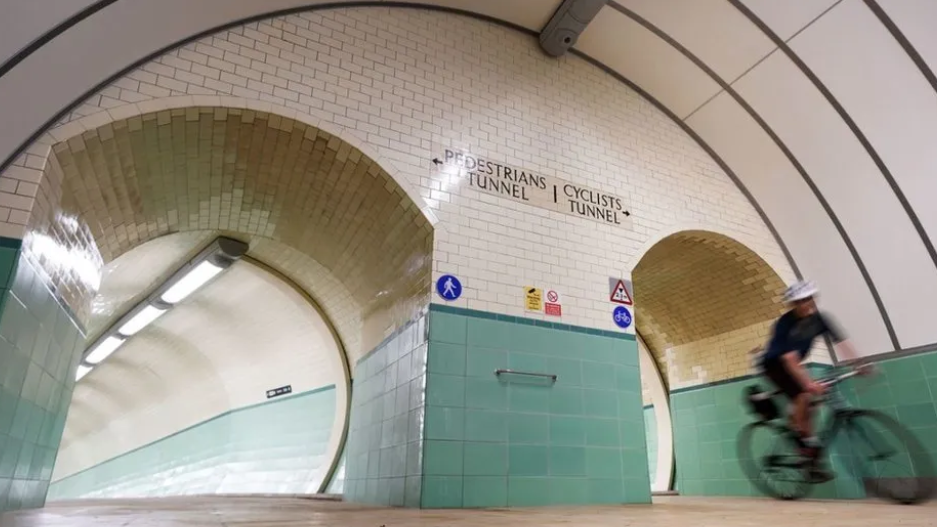 Two tunnels covered in green and cream tiles. There is a sign pointing to one which reads 'pedestrian tunnel' and one pointing to the other saying 'cyclists tunnel'. A man on a bike, dressed in black and a helmet is coming out of the cyclists tunnel.