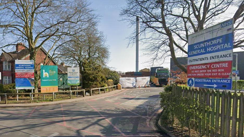 The entrance to a hospital with fencing either side of a road going into it. Either side of the fencing you can see signs saying 'NHS Scunthorpe General Hospital, Outpatients Entrance, Emergency Centre'.