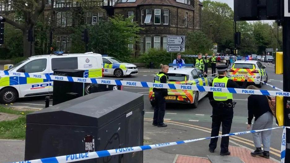 A police cordon in place at a busy junction. Present are several uniformed officers standing on the street corner, with four police vehicles around them. A roadsign, pedestrian crossing and other street furniture can be seen.