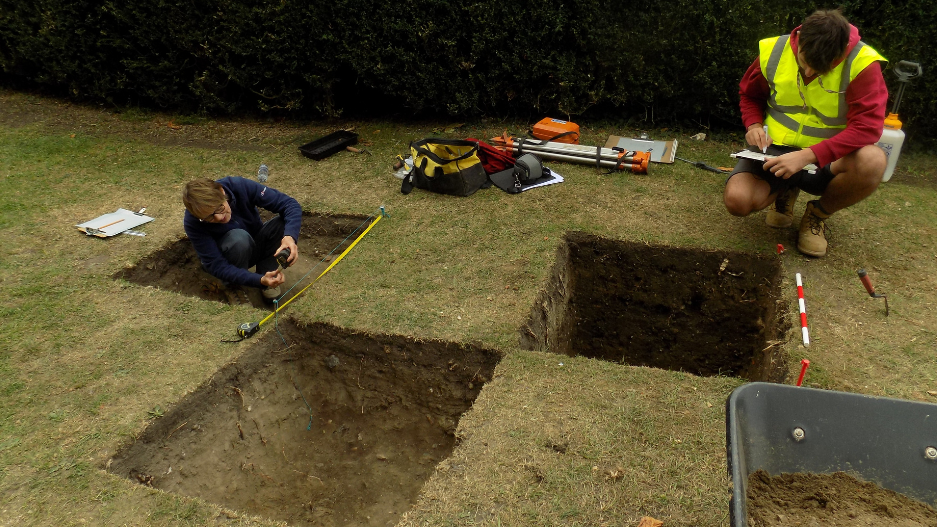 An archaeologist squatting in a rectangular shaped pit making measurements. Another, wearing a high -vis vest, making notes on a clipboard next to two further rectangular shaped pits.