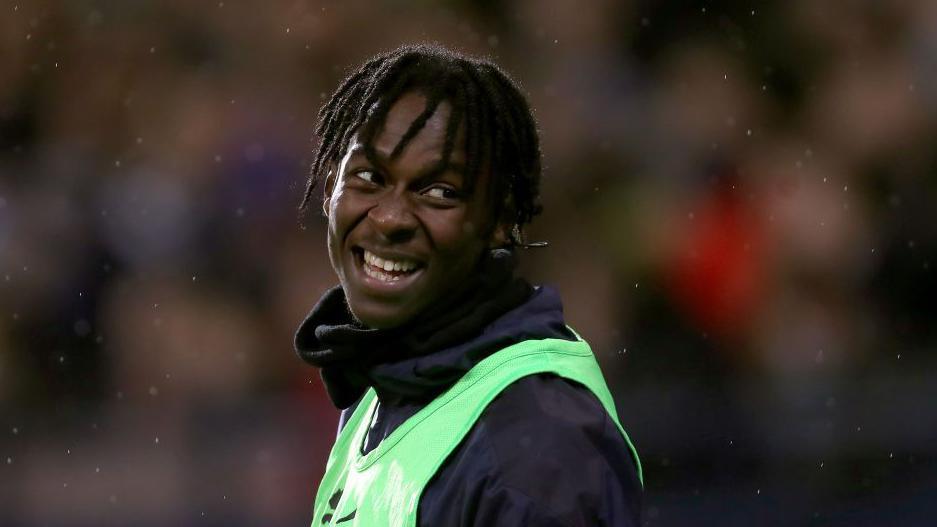 Quevin Castro is smiling as he looks over his shoulder. He has short dreadlocks and is wearing a tracksuit top and green bib.