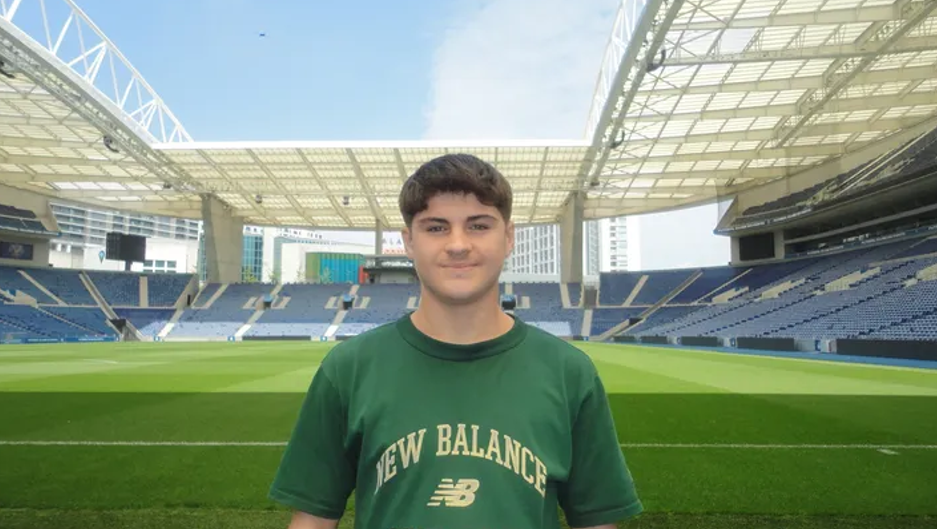 A young man with brown hair, wearing a green t shirt with the words NEW BALANCE written across the front in beige writing. He is smiling and looking at the camera and is stood with a football pitch and stands behind him.