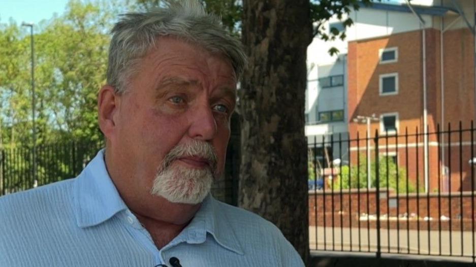 A man with short grey hair and a beard wearing a blue shirt.
