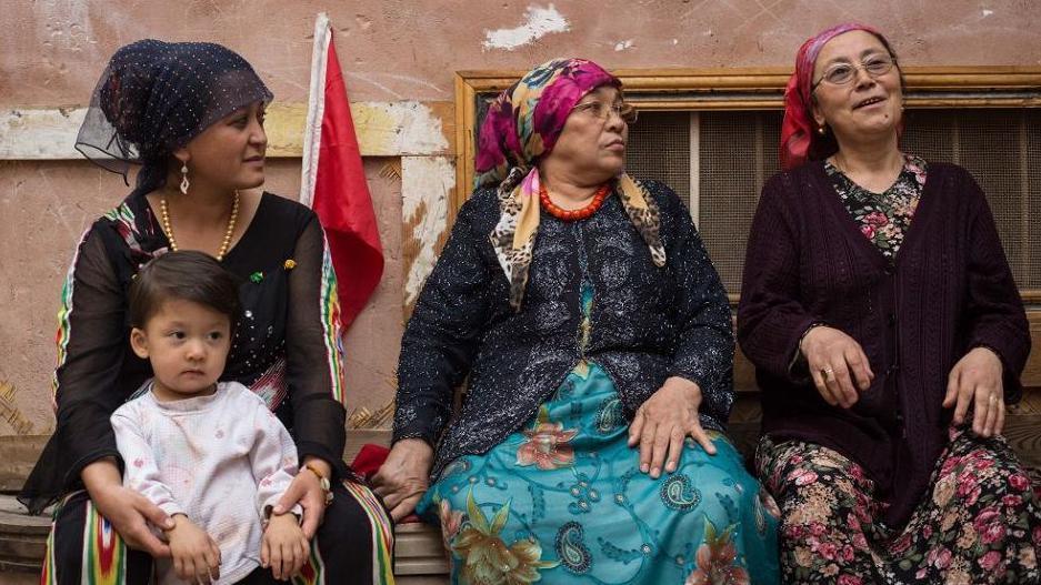 A young Uyghur women,  with a small child on her knee, sits next to two older women on a street in Kashgar City, Xinjiang. All three are dressed in traditional garments with head scarves