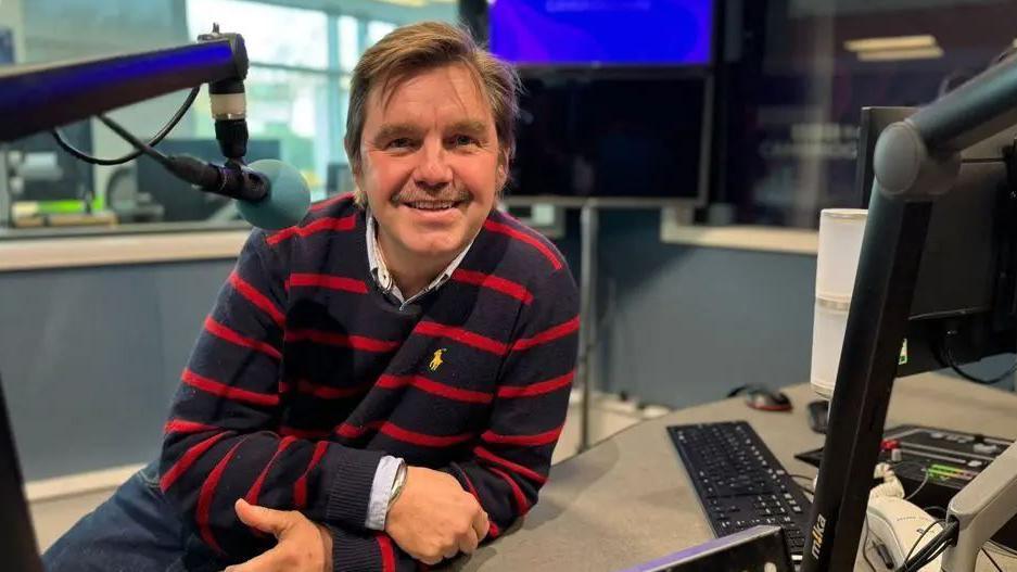 Nik Johnson wearing a black-and-red striped jumper sitting in a radio studio. He is leaning on a table in front a microphone and is smiling at the camera.