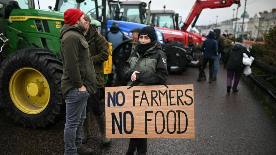 A female dressed in a green and black rain coat holds a sign saying No Farmers No Food. A crowd of people and Green, red and blue tractors are in the background. 