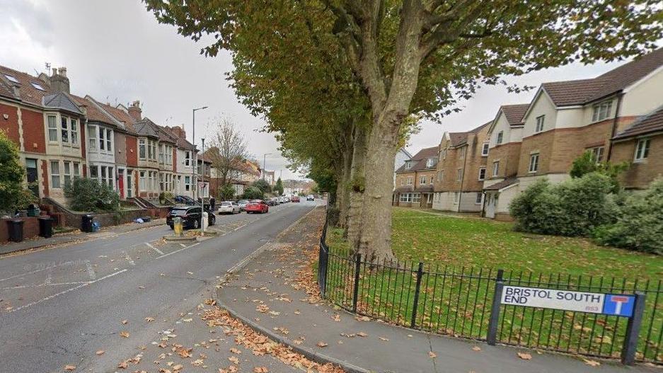 A Google street view of the junction of St John's Lane and Bristol South End, with houses and cars on either side of the main road and a row of large trees