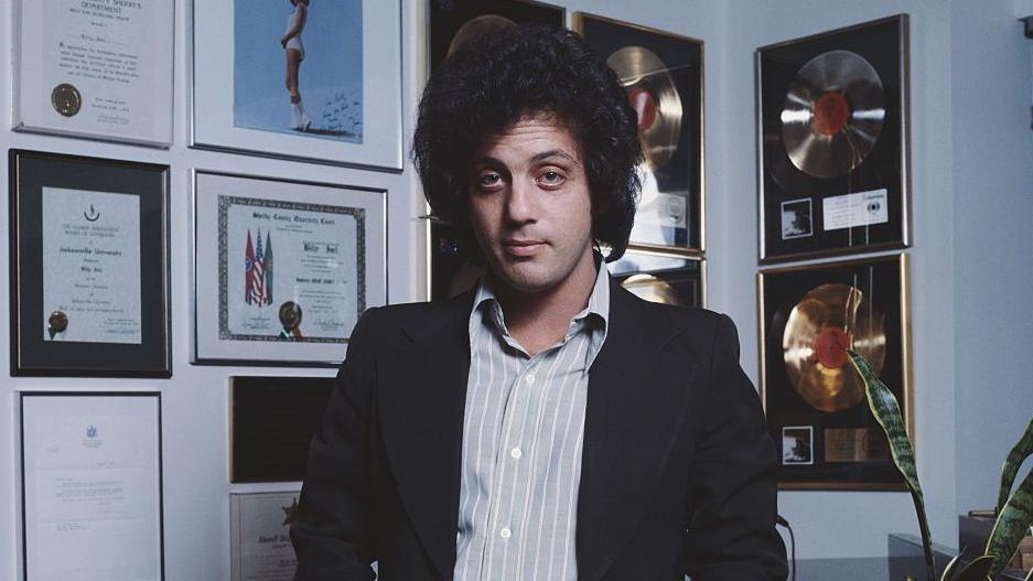 A man is looking directly at the camera, with a lot of frizzy dark hair. He is wearing a blue striped button down shirt with a black jacked, and he's standing in front of a wall of framed awards.