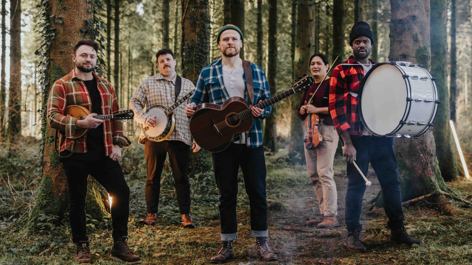 5 members of Rend Collective standing in a forest. From the left, Wil Pearce has short dark hair and a beard and is holding a ukulele. Next to him, Stephen Mitchell has short, curly black hair and a moustache, and holds a banjo. In the middle, Chris Llewellyn has a beard and a green beanie, and holds a guitar. Next to the right is Jonathan Chu, he has long dark hair and is holding a violin. Next is Daniel Jones wearing a black beanie and holding a large drum strapped around his neck