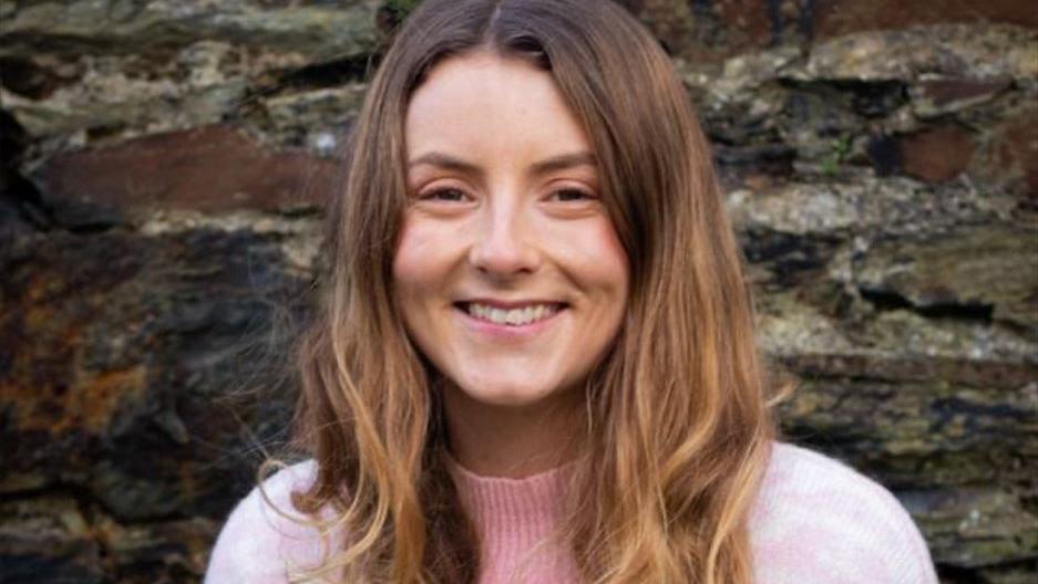 Meghan Kennish has long light brown hair worn down, and is smiling, wearing a light pink jumper, and standing in front of a stone wall.