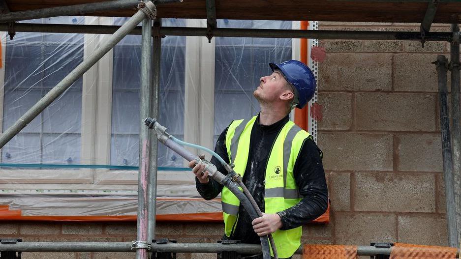 A construction worker wearing a blue helmet and a yellow high-viz jacket works on scaffolding outside a building.