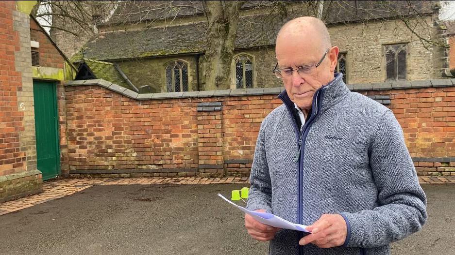 Crooks reading a document standing in the courtyard of the school hall with a church in the background