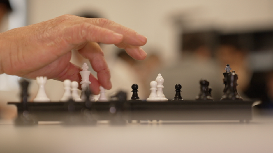 A zoomed view of a chess game. A hand is moving a white pawn towards the opposing black pieces. The rest of the view is blurred.