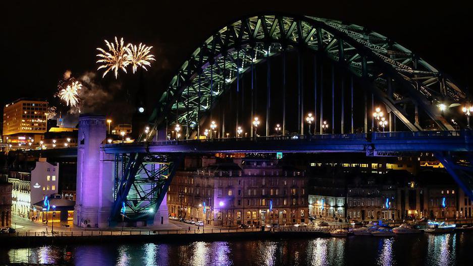 Fireworks exploding in the night sky, next to the Tyne Bridge and above Newcastle quayside, which is illuminated by streetlights. 